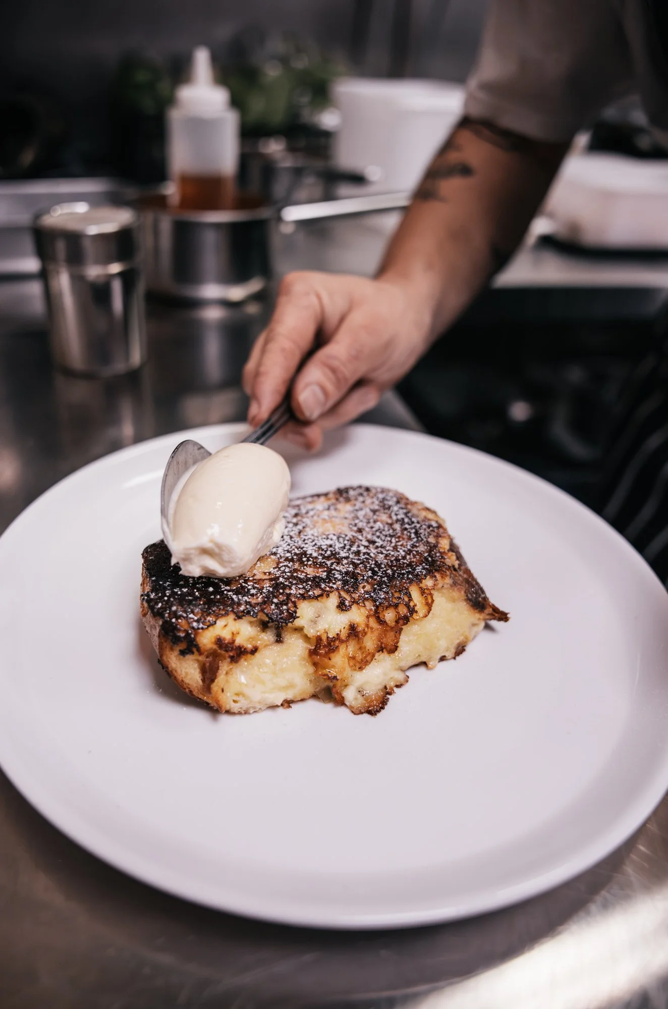 A slice of French toast with powdered sugar, topped with a scoop of vanilla ice cream on a white plate, in a restaurant kitchen.