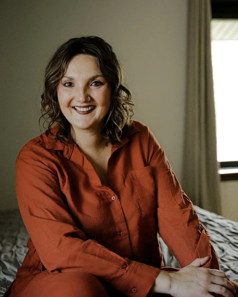 A woman with dark curly hair smiling, wearing a rust-colored shirt, sitting on a bed in a room with beige walls and a window with curtains.