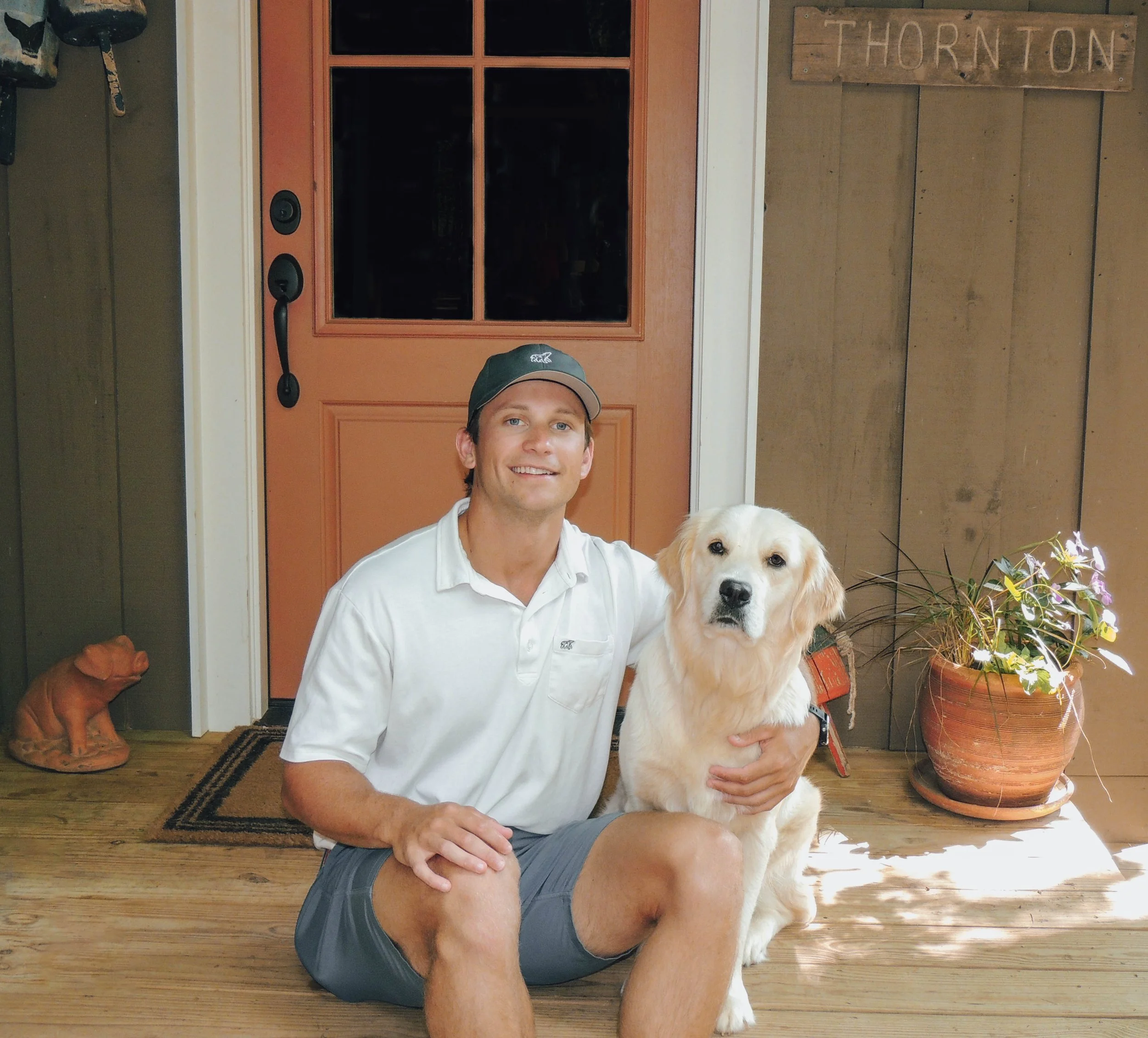 A man sitting on a wooden floor, smiling, with a golden retriever puppy. Both are in front of a front door with a wooden sign that reads "Thornton." There are plants in a large pot to the right and a person’s leg and foot visible in the background on the left.