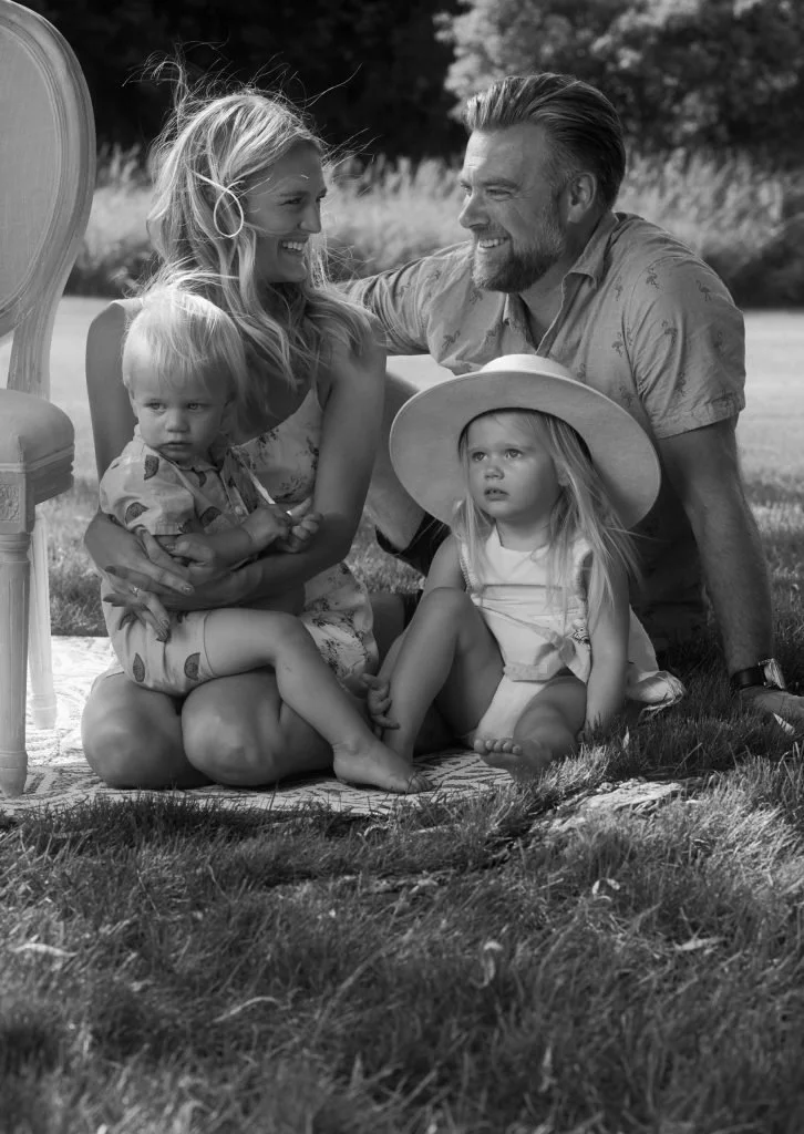 A family of four sitting outdoors on a blanket; a woman holding a young boy, a man smiling, and a young girl with a large sun hat sits nearby, in a grassy area with trees in the background.