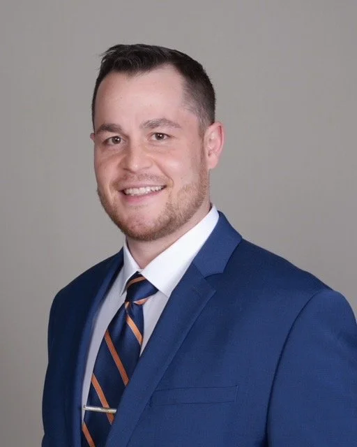 Young man wearing a blue suit, white shirt, and a striped tie, smiling against a plain background.