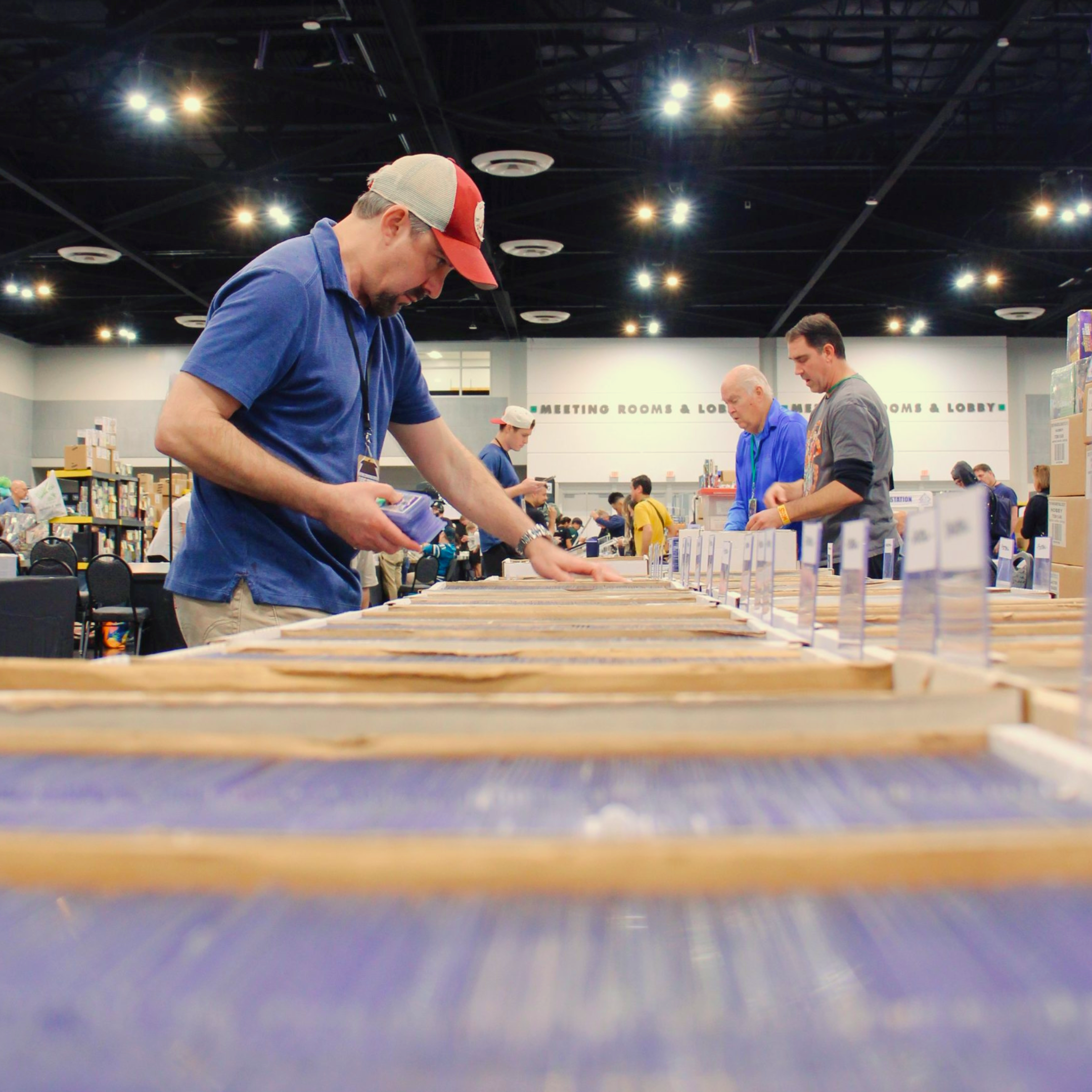 People browsing books at a convention or event in a large indoor space with bright ceiling lights and sign reading 'Meeting Rooms & Lounge' in the background.