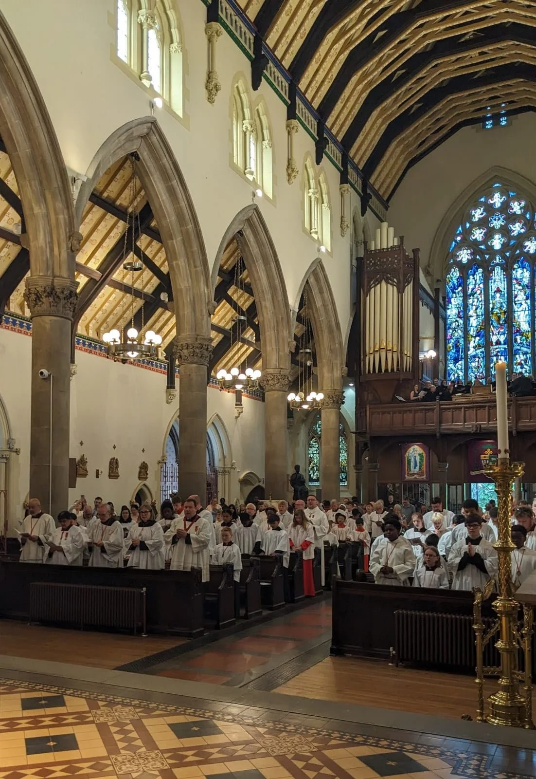 Congregation of people dressed in white robes inside a church, standing at pews during a religious service, with stained glass windows and wooden architecture in the background.