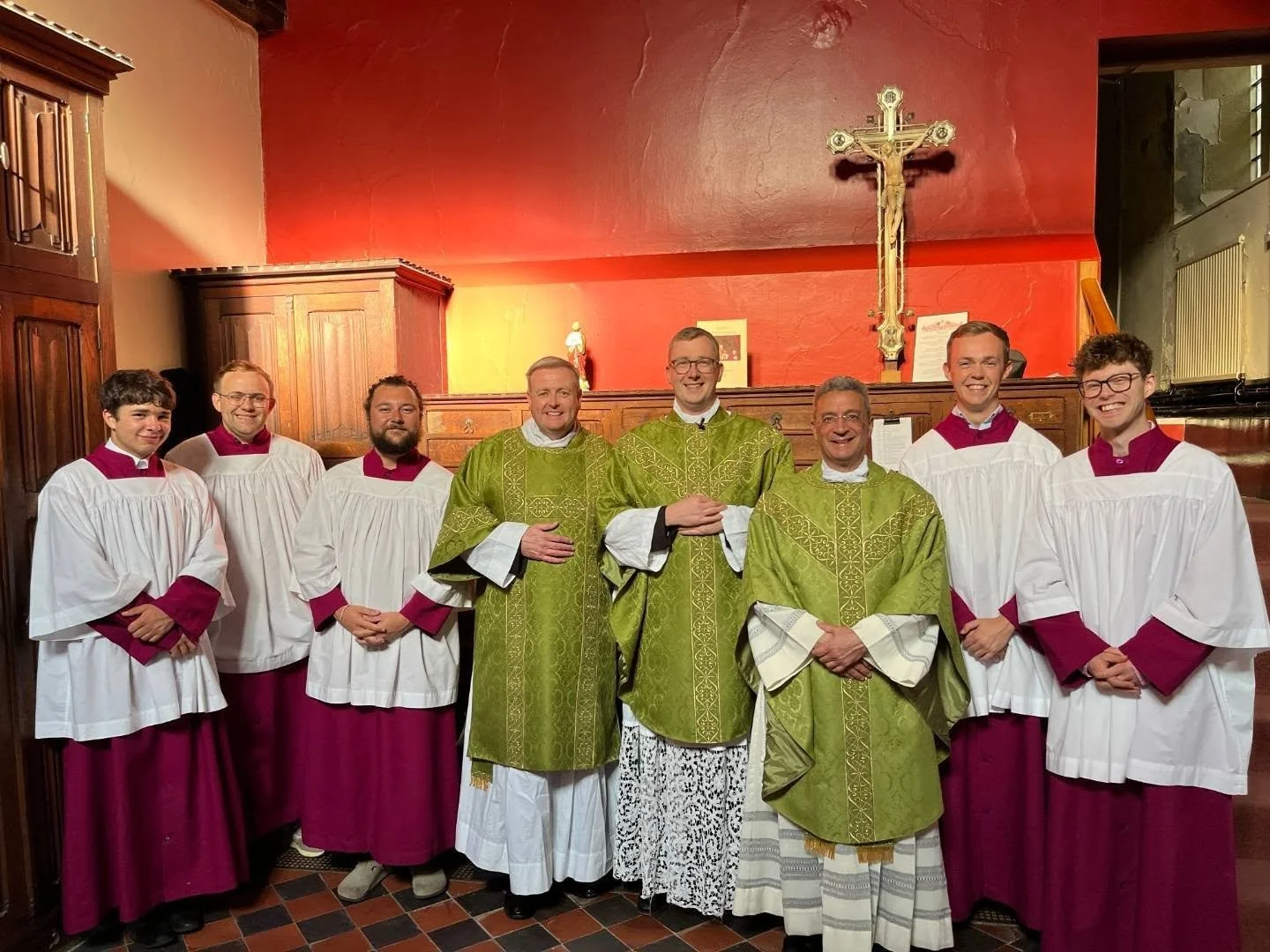 Group of nine priests standing in church, some with arms crossed, smiling, in front of a red wall with a large crucifix and religious decorations.
