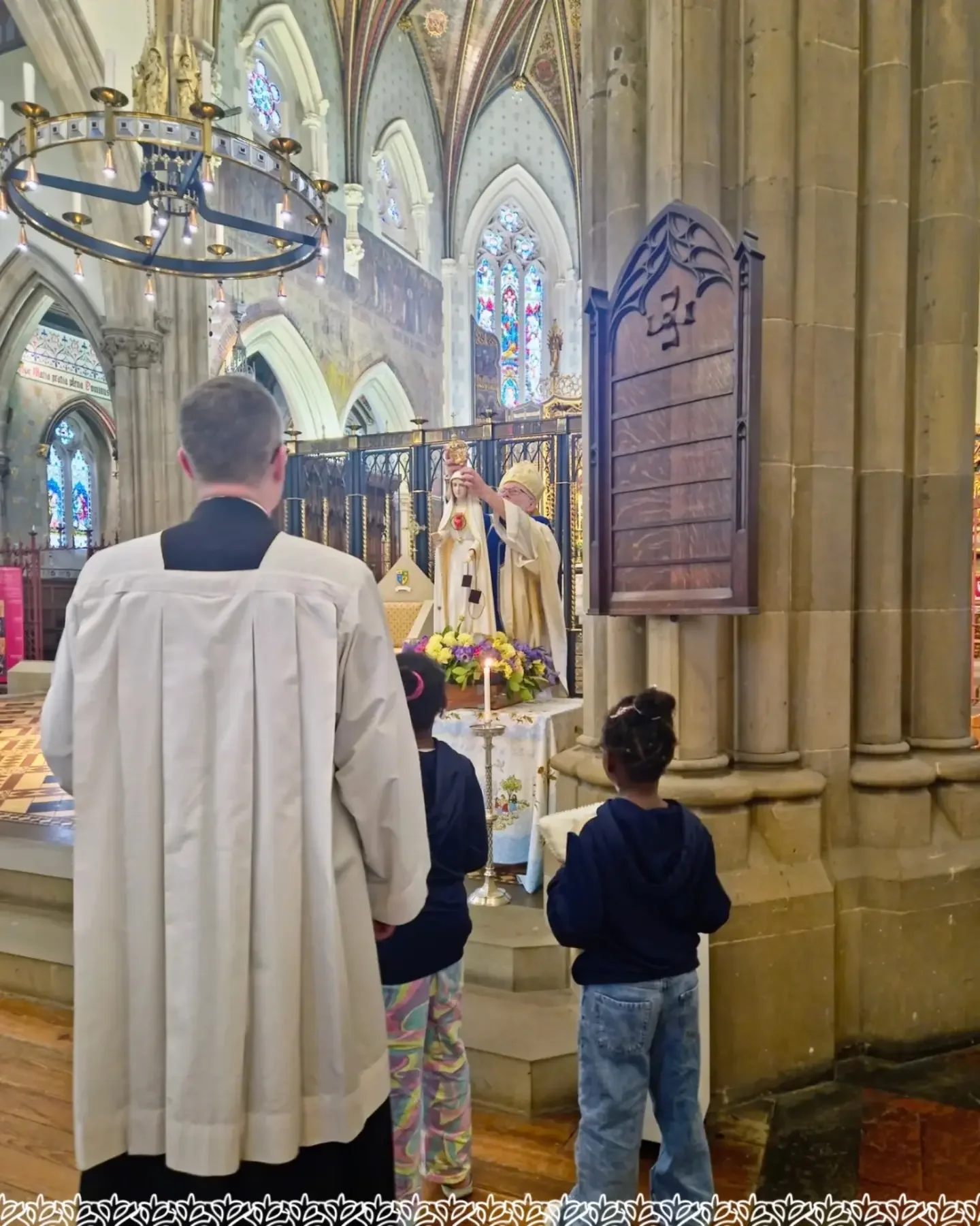 Inside a Gothic-style church with stained glass windows, a priest or pastor performing a religious ceremony at an altar, with children and a man in choir robes participating or observing.