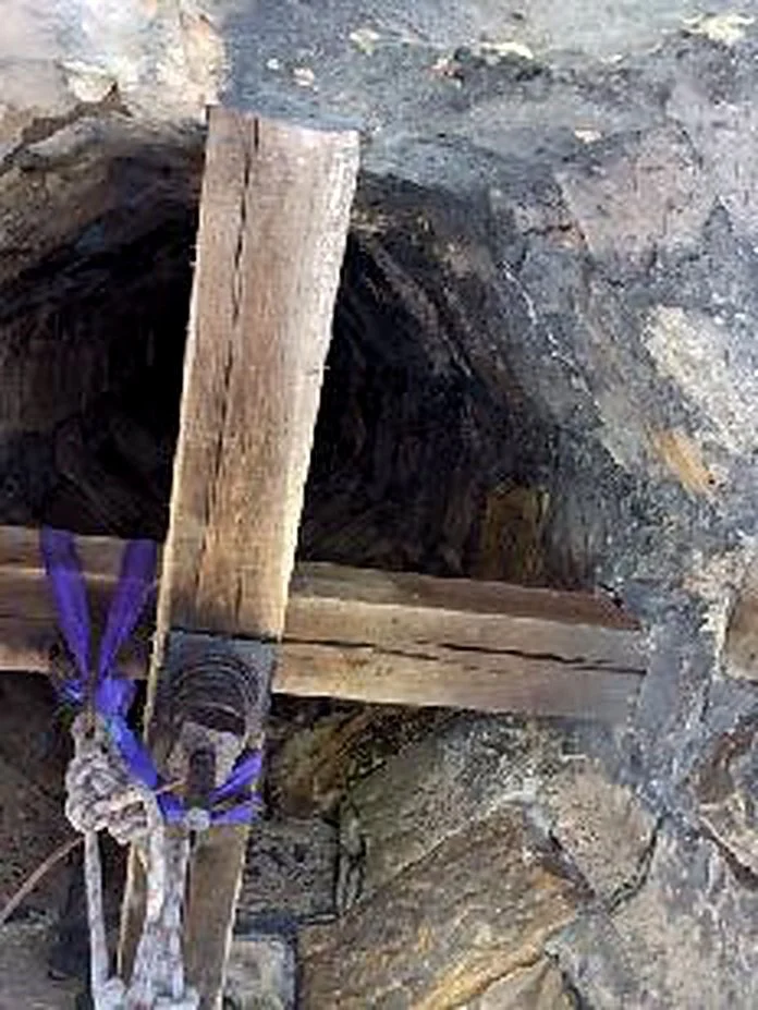 A makeshift wooden cross tied with purple and white fabric against a stone wall.