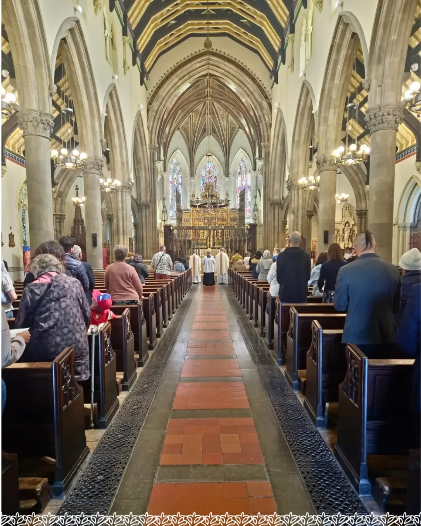 Inside a large church, people are gathered for a service. The church has tall stone columns, high vaulted ceilings, and stained glass windows at the front. The aisle is lined with wooden pews, and there are clergy members in white robes at the altar.