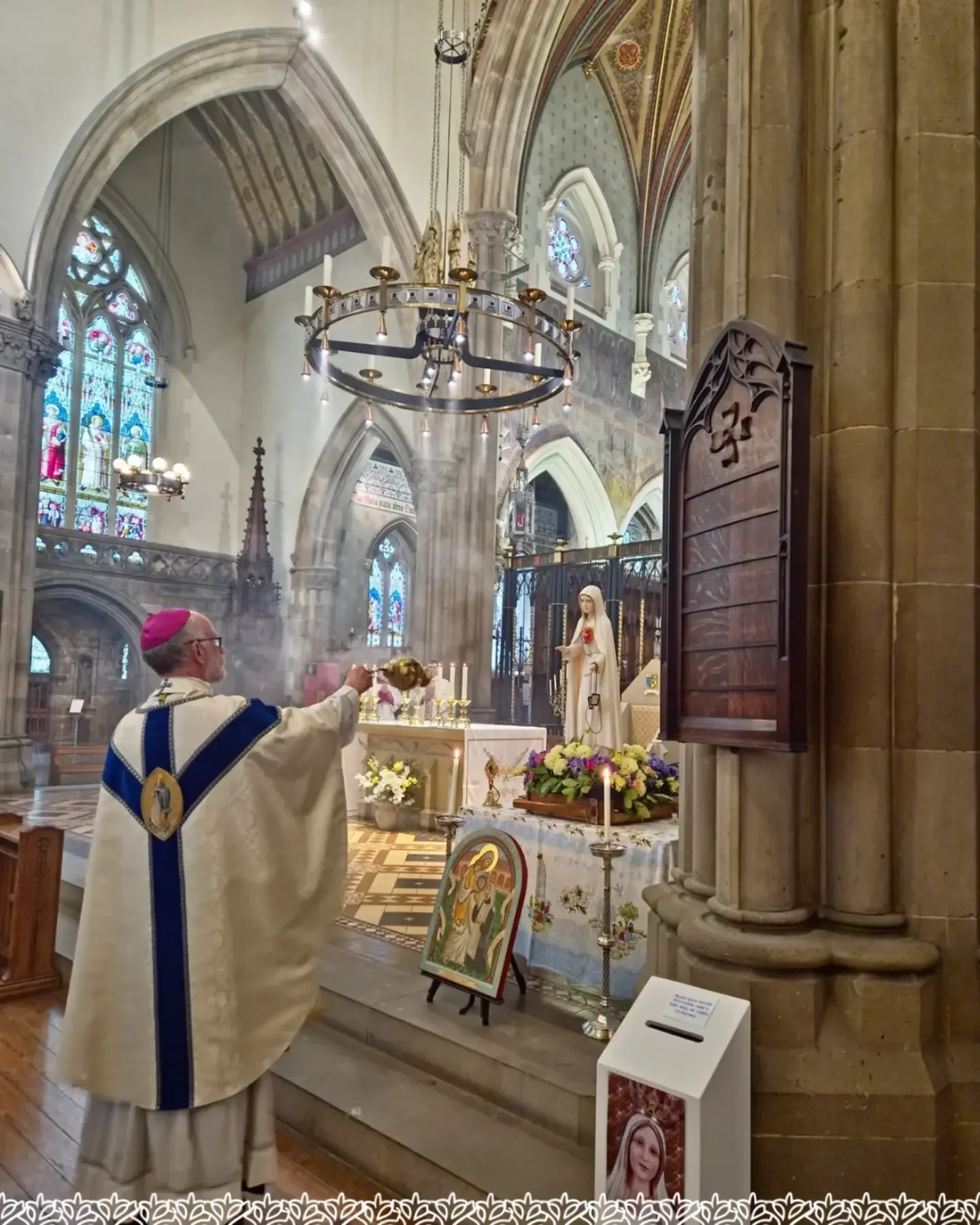 A priest in white and blue vestments performing a religious ritual at an altar inside a church. The church has stained glass windows, chandeliers, and religious statues.