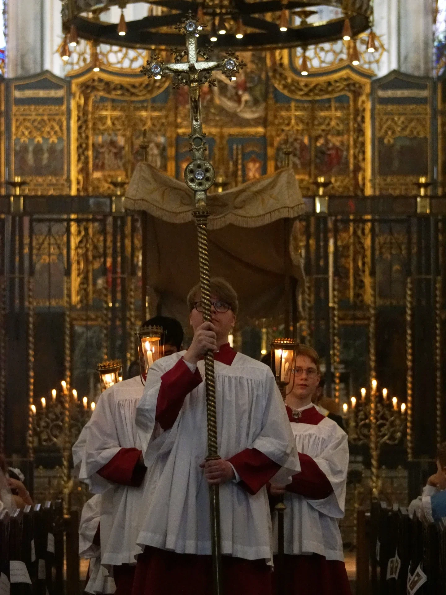 Young people in white robes and red sleeves participate in a religious procession inside a church, carrying candles and a cross. The church interior features ornate gold decorations and stained glass windows.