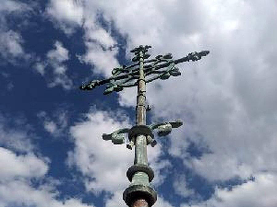 A tall metal cross with ornate details standing against a background of blue sky and white clouds.