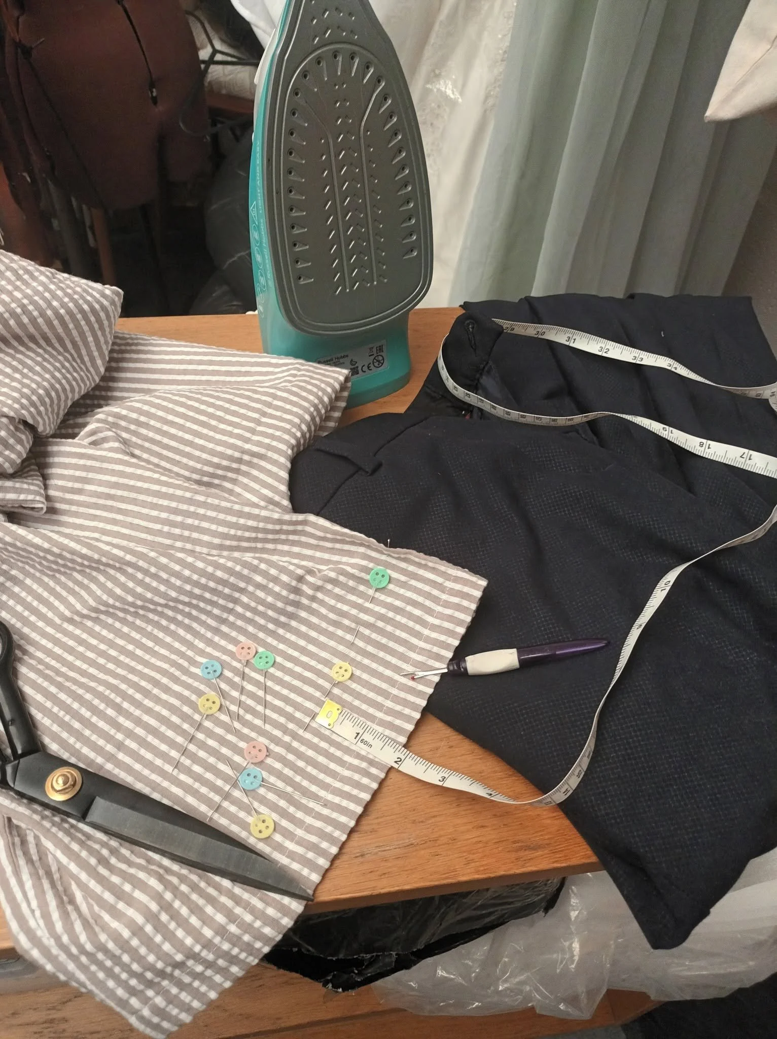 A sewing workspace with various sewing tools and supplies on a wooden table. There is a striped fabric, a collection of colorful buttons, a measuring tape, and part of a sewing machine. An iron is also visible in the background.