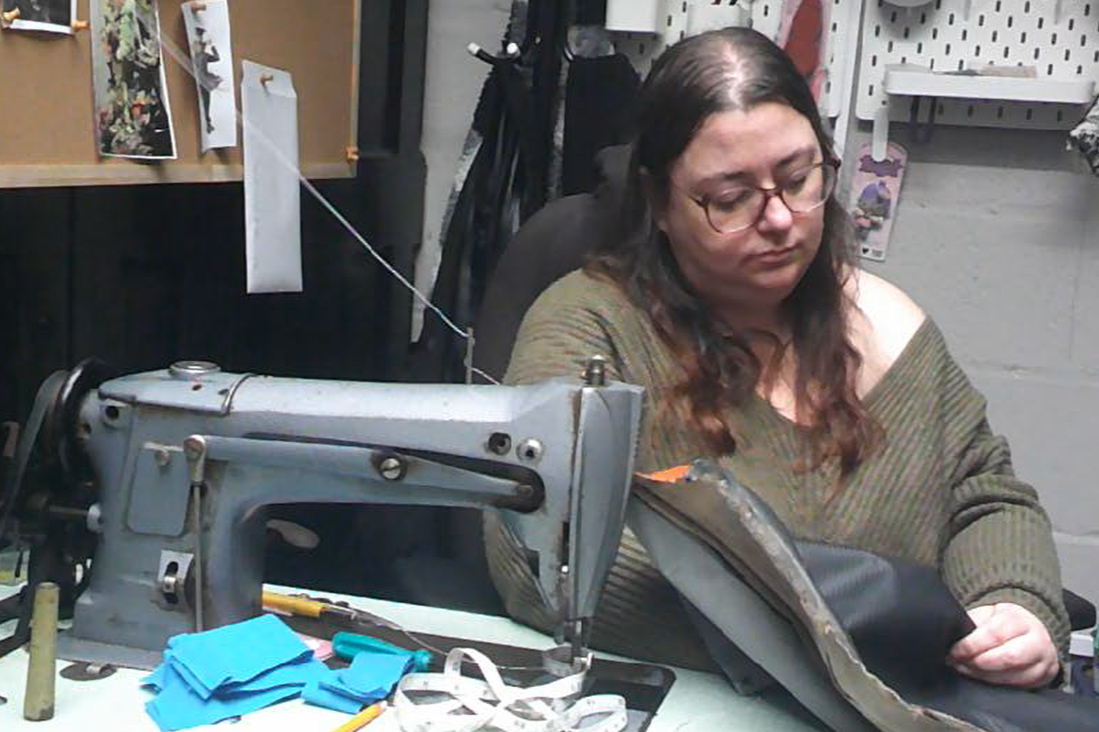 A woman with glasses and long brown hair working on a black leather handbag at a sewing machine in a workshop.