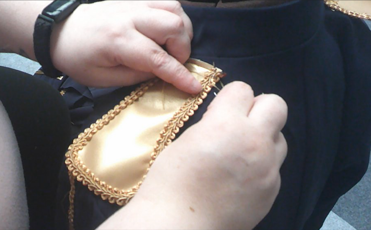 Close-up of a person's hands sewing a gold-trimmed gold fabric onto a dark fabric, possibly part of a costume or uniform.