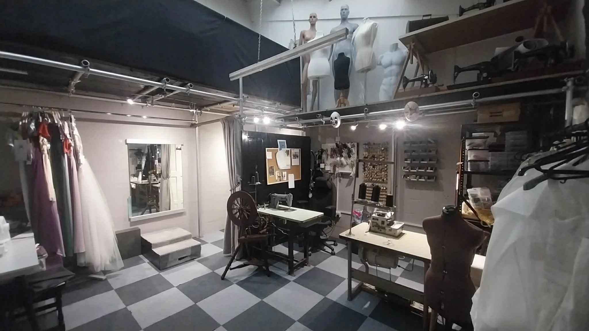 A sewing studio with dress mannequins, fabric dresses, sewing machines, and fabric rolls, featuring a black and white checkered floor.