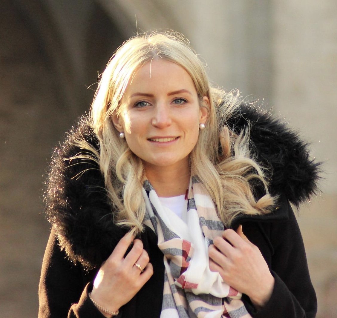 Nahaufnahme eines lächelnden weißen Frauen mit blonden, lockigen Haaren, die eine schwarze Jacke mit Fellkragen trägt.