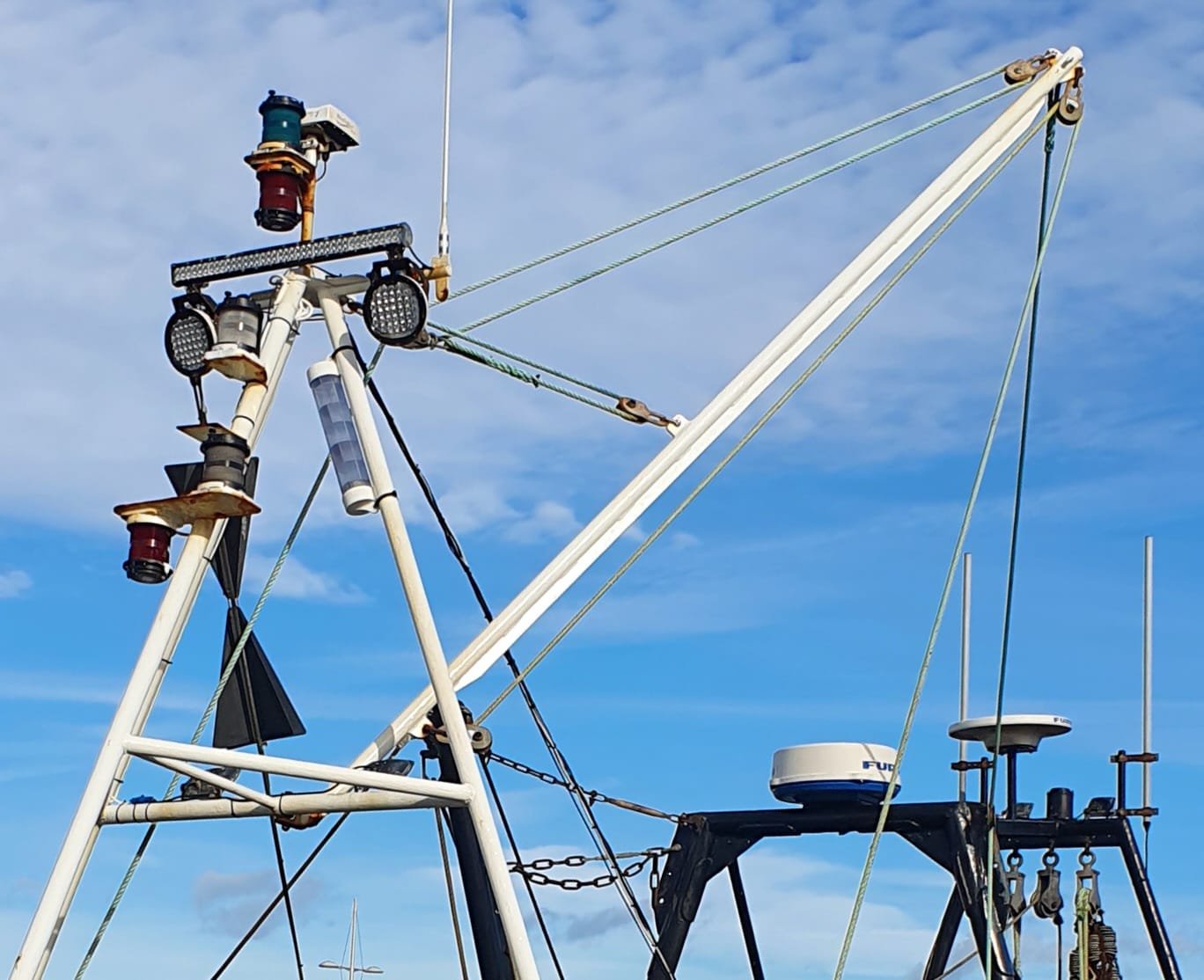 Close up of a fishing vessel's mast fittings, lights and rigging equipment against a partly cloudy sky. imagery from the chandlery.