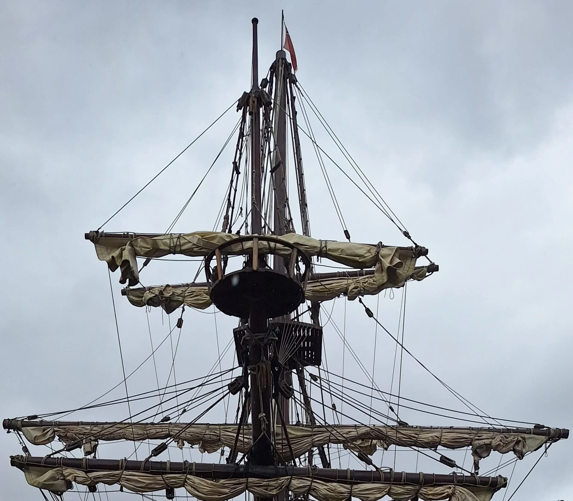 Bow of a traditional tall ship, mast, rigging and sails set against a cloudy sky. Imagery from The Log