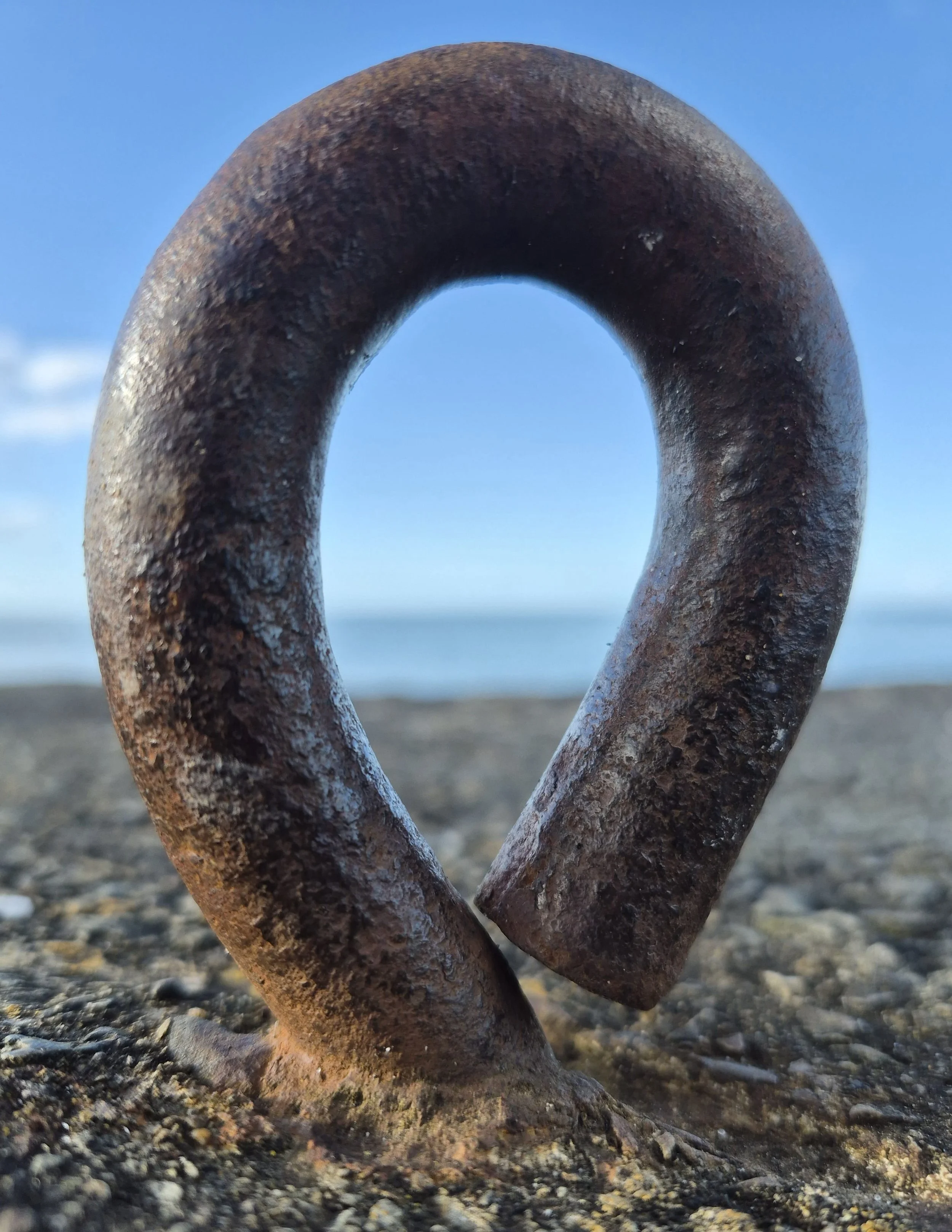 Weathered metal mooring ring set in gravel and sand, ocean visible in the blurred background. Texture imagery for AbeySea's maritime themes