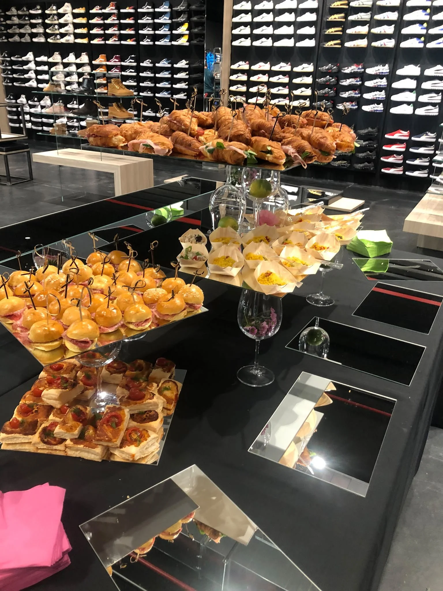 Esposizione di snack e finger food, tra cui mini panini, croissant ripieni e altri snack, in un negozio con pareti ricoperte di scarpe sportive.