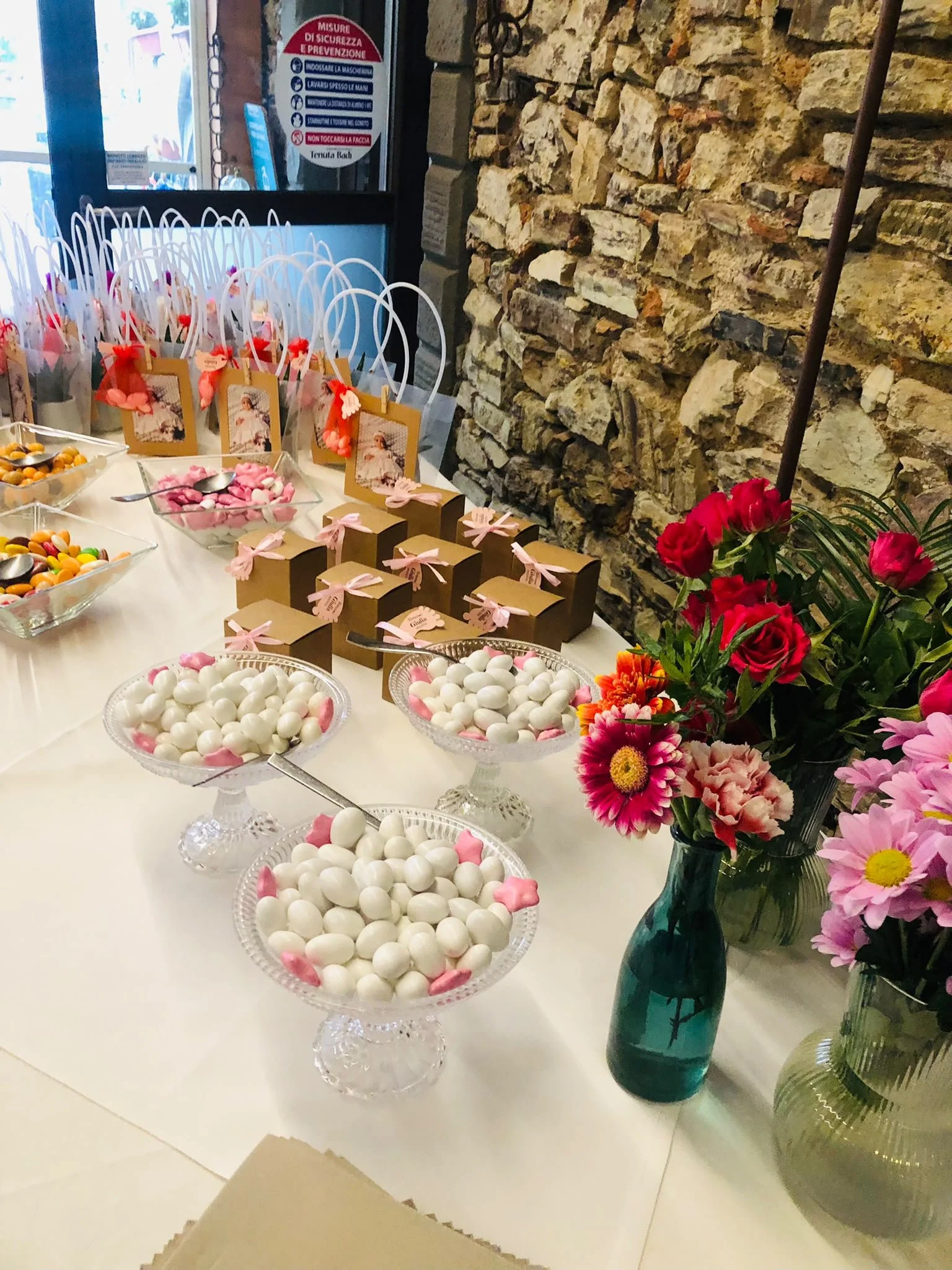 Tavolo decorato con caramelle colorate, cioccolatini e un mazzo di fiori in un vaso verde, con uno sfondo di parete in pietra rustica