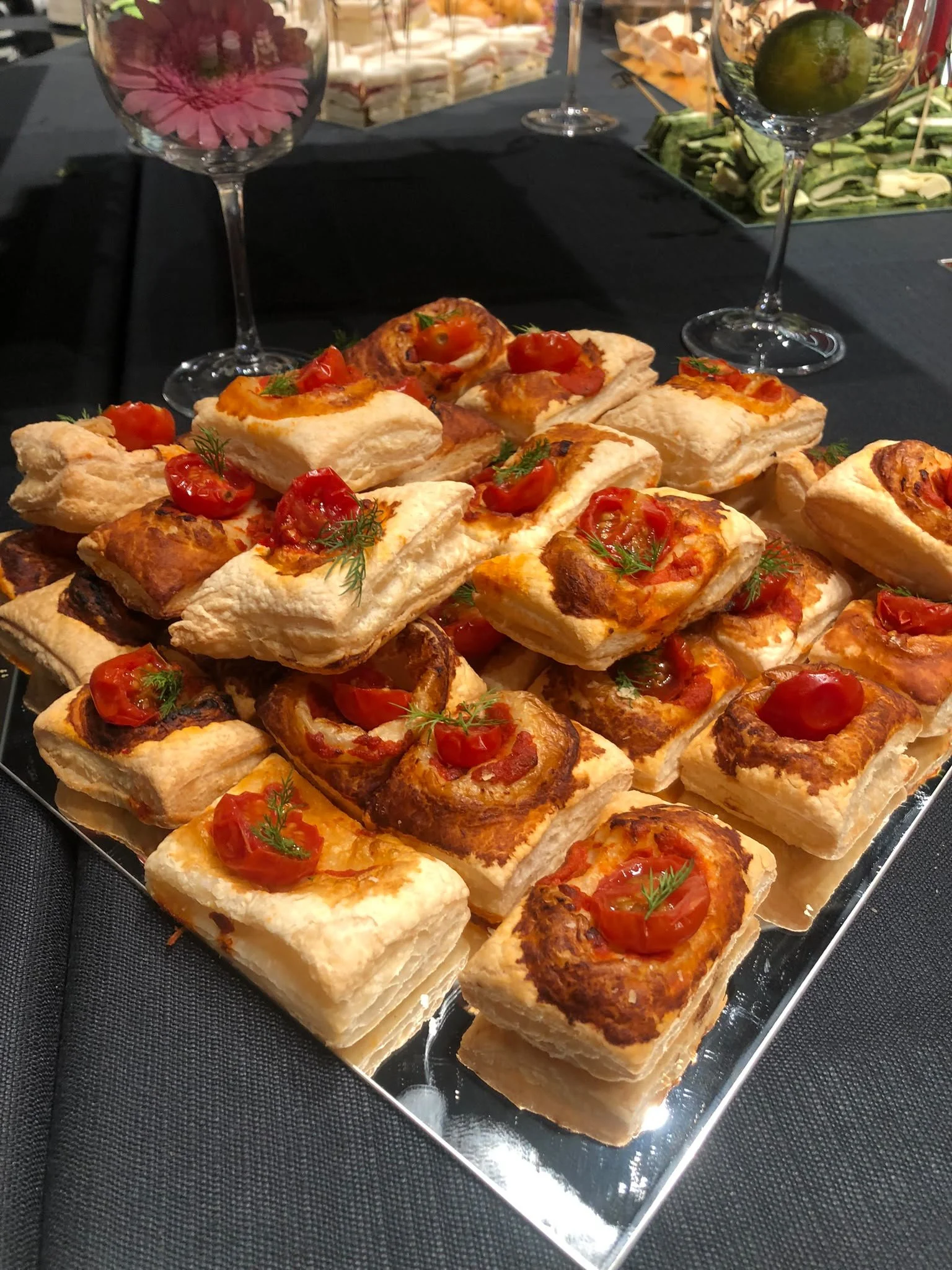 Selezione di antipasti caldi e freddi, tra cui mini croissant salati con pomodorini e erbe aromatiche, serviti su un vassoio di specchio, con calici di vino.