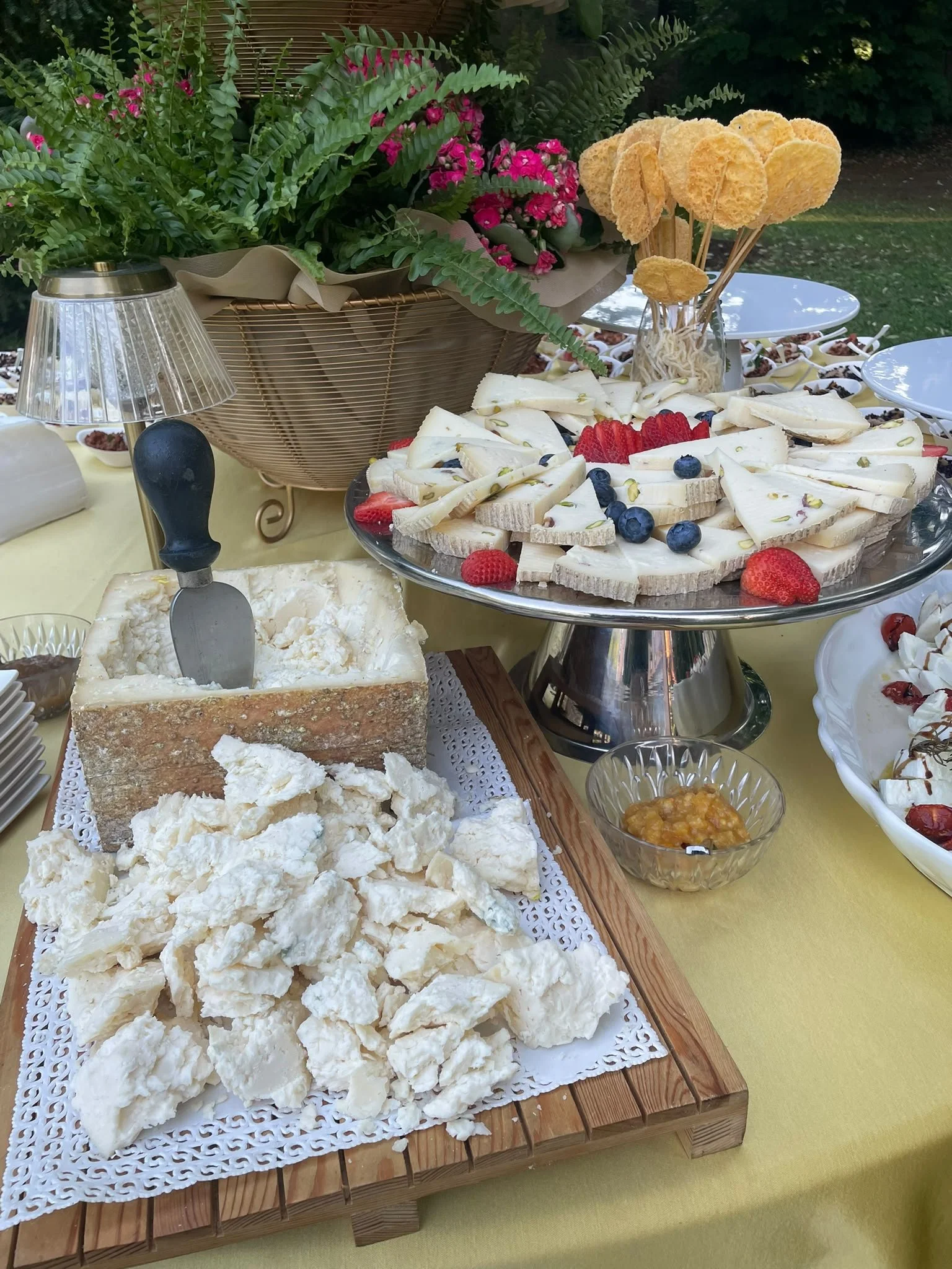 Un buffet di formaggi con frutta fresca come fragole e mirtilli, decorato con fiori colorati e un vaso con piante verdi. Sono presenti anche dei crostini di pane e altri dolci e snacks in ciotole.