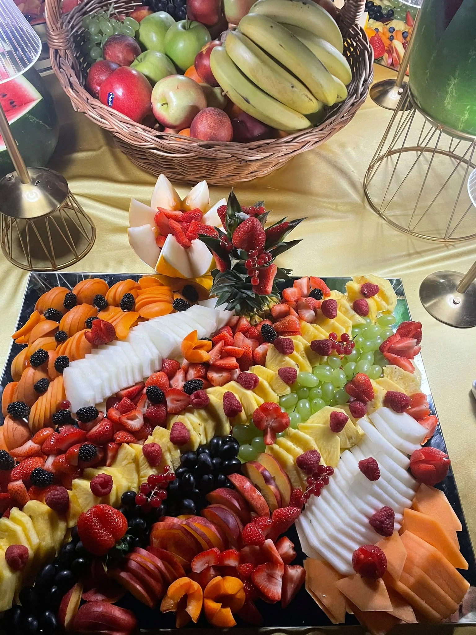 Vassoio di frutta fresca tagliata con melone, ananas, fragole, uva verde e nera, lamponi, mirtilli, pesche e fetta di anguria. Sopra, una ciotola di frutta a forma di fiore decorativa e una coppa di frutta con o altri frutti.