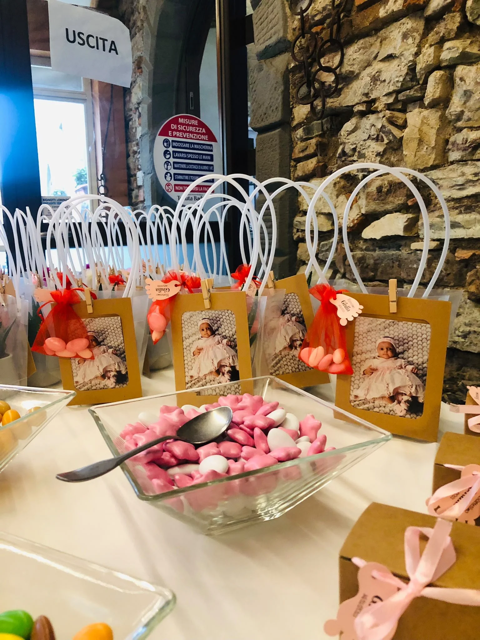 Decorazioni e dolcetti per un battesimo, con cartellini con fotografie di un bambina e sacchetti di confetti rosa e rosso, in un ambiente rustico ed elegante.