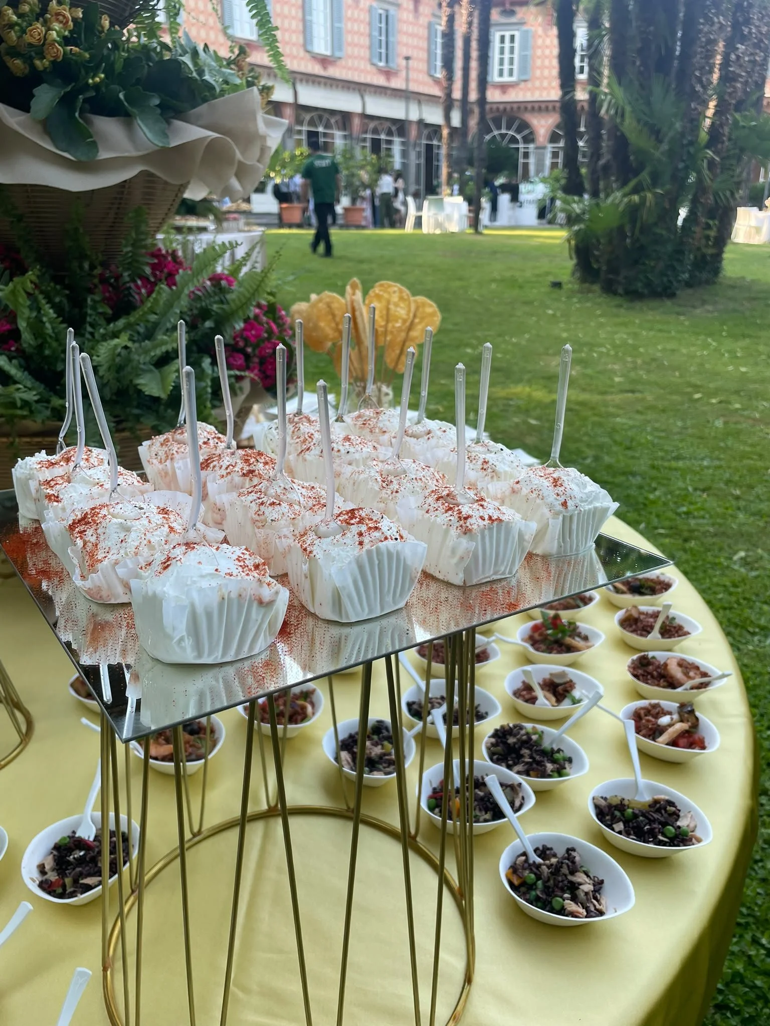 Una tavola elegante con monoporzioni di ricotta dolce e paprika affumicata sopra, serviti in coppe di carta, e ciotole di vari primi freddi, in uno spazio all'aperto con erba, palme e un edificio storico sullo sfondo.