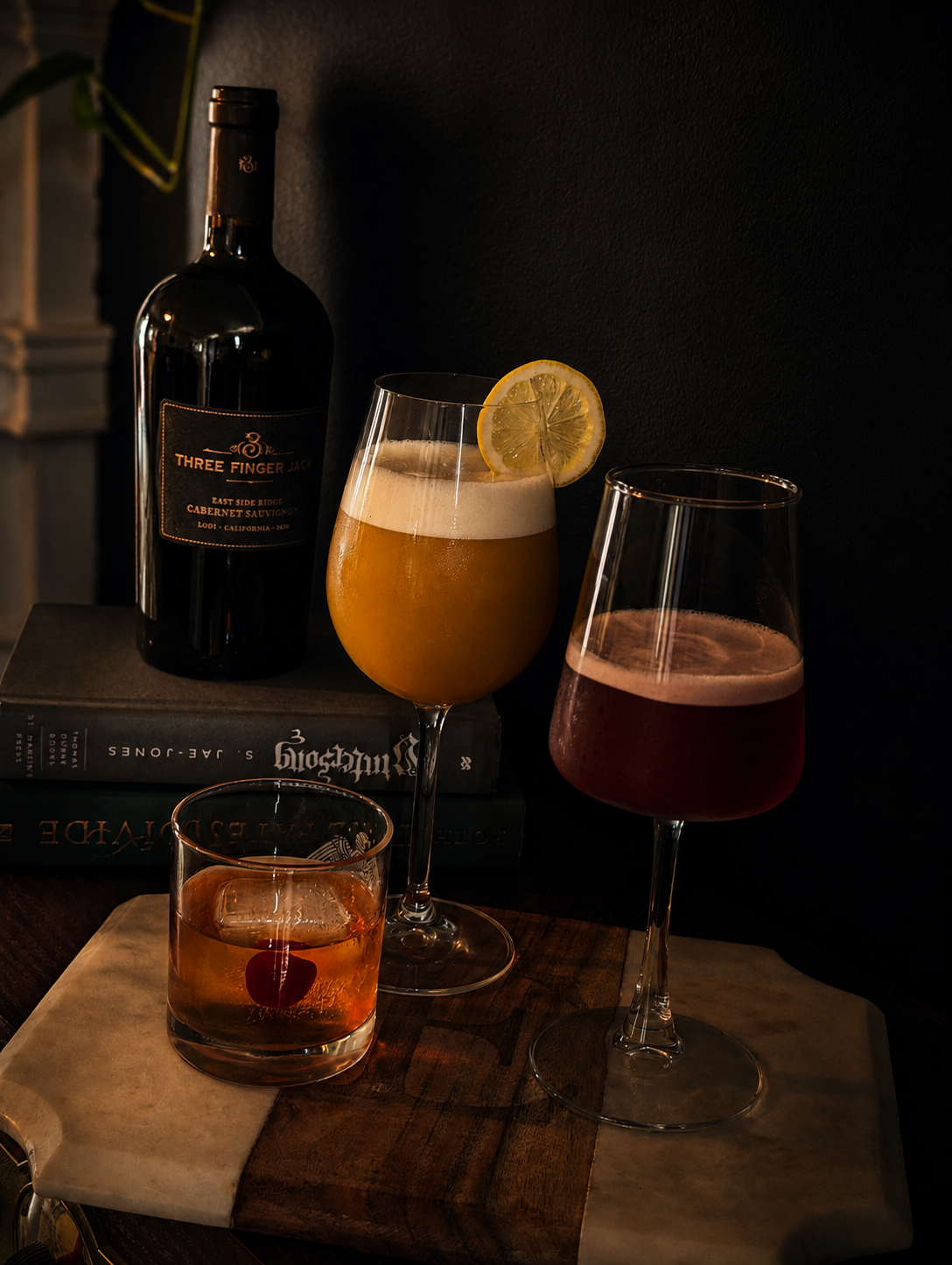 Three cocktails on a wooden surface, with a bottle of wine and books in the background. The middle cocktail has a lemon slice garnish, and another has a cherry inside.