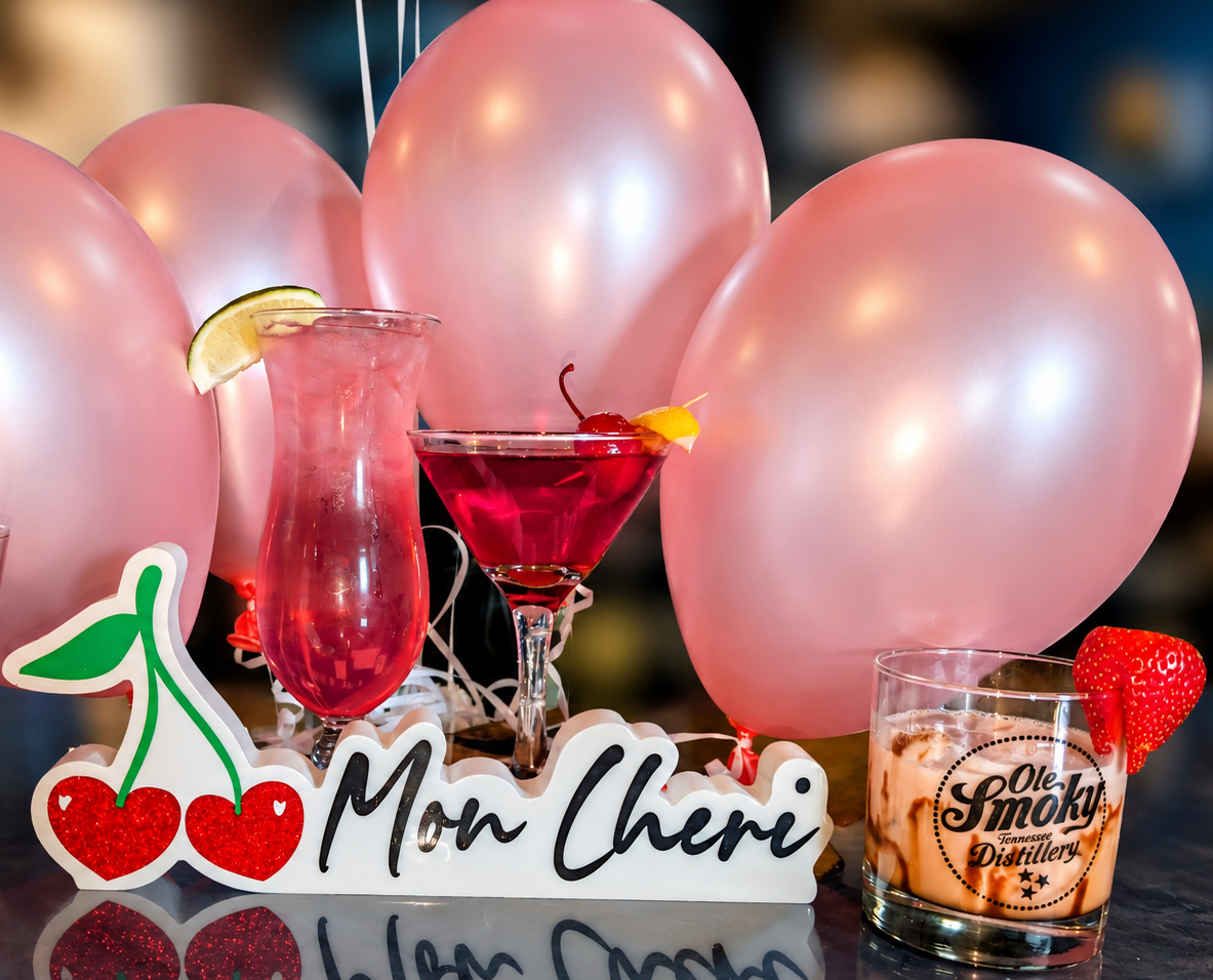 A celebration setup with pink balloons, cherry and lemon garnished drinks including a vintage pink cocktail, a martini with a cherry and lemon twist, and a glass of whiskey with a strawberry, all arranged on a reflective surface with a decorated sign reading 'Mon Cheri' and a cherry sticker.