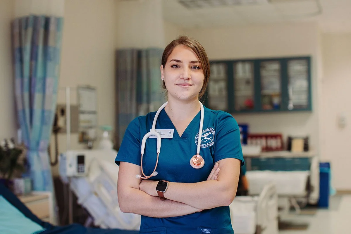 A young female nurse standing with arms crossed in a hospital room. She is wearing blue scrubs and has a stethoscope around her neck, looking confidently at the camera.