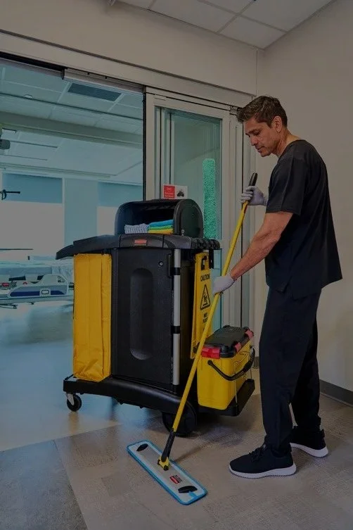 A male hospital staff member mopping the floor with a mop and bucket, wearing gloves, near a cleaning cart in a hospital corridor