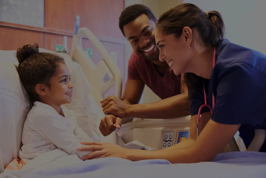 A young girl in a hospital bed smiling at healthcare providers. Two medical staff, a nurse and a male doctor, are engaging with her, holding her hand and smiling.