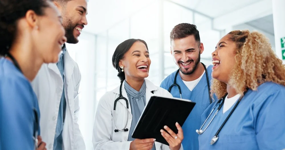 A diverse group of five healthcare professionals, including doctors and nurses, are gathered in a bright, modern medical facility, smiling and laughing while looking at a tablet device.