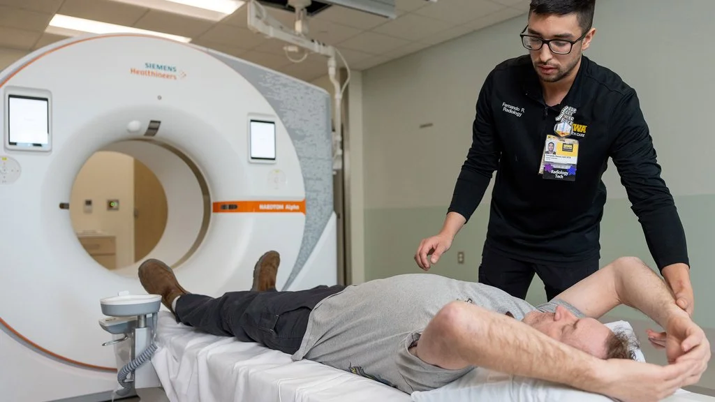 A male patient lying on a hospital bed with his arms behind his head, about to undergo a CT scan in a radiology department. A radiology technician stands nearby, positioning the patient.
