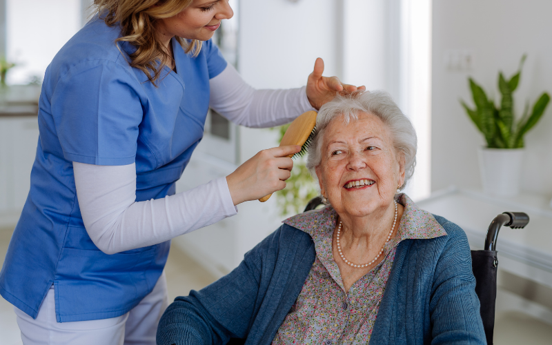 A caregiver brushing the hair of an elderly woman sitting in a wheelchair, smiling in a bright room.