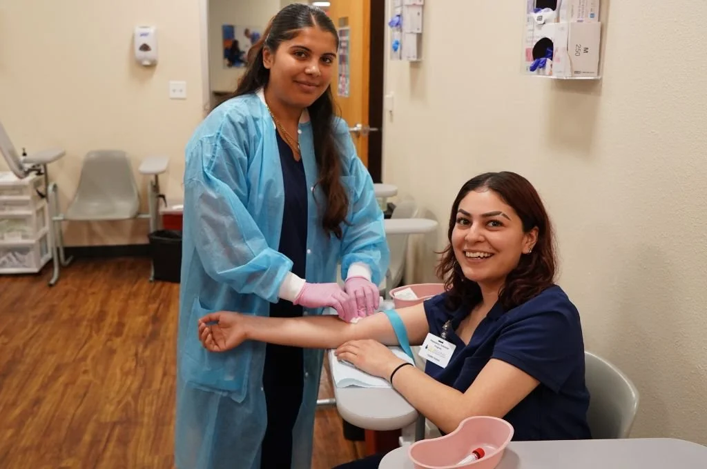 A woman in a dark blue uniform sitting in a medical clinic, with her arm extended, smiling at the camera while a healthcare worker in a blue gown and pink gloves prepares to draw blood or give an injection.