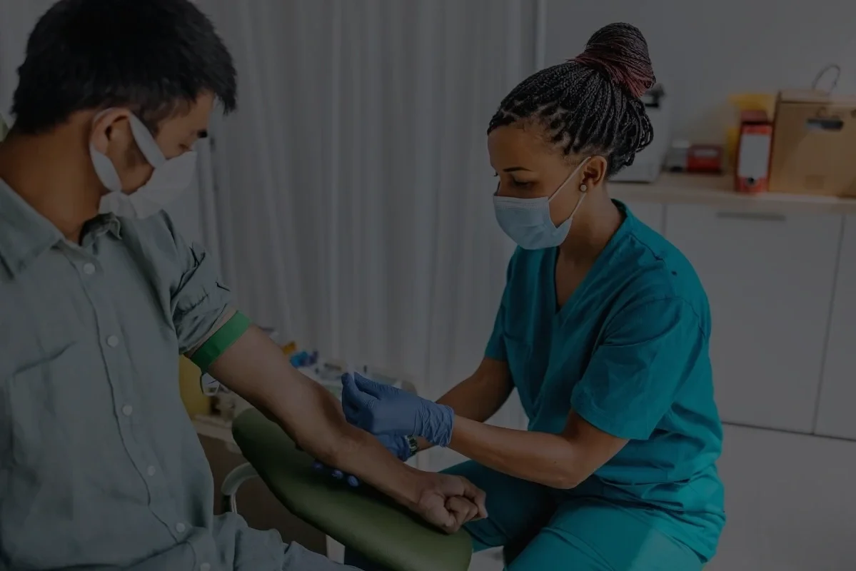 A healthcare worker in scrubs and mask prepares to draw blood from a patient's arm. The patient is seated with their arm extended, also wearing a mask.