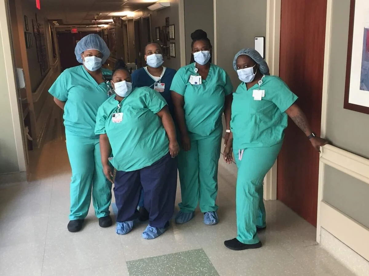 Group of five healthcare workers wearing green scrubs and face masks standing in hospital corridor.