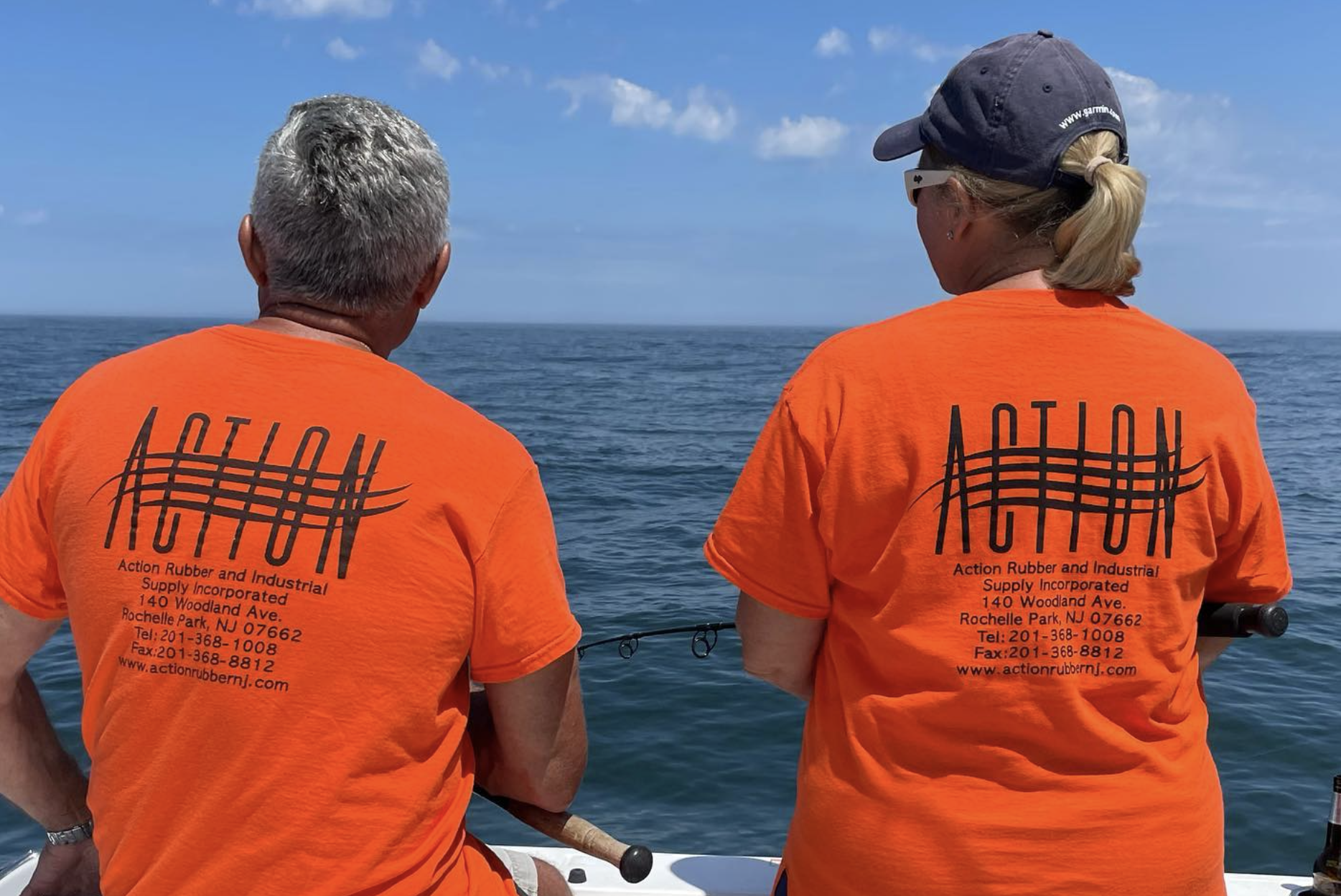 Two people wearing orange t-shirts with 'ACTION' printed on the back, standing on a boat with the ocean and blue sky in the background.