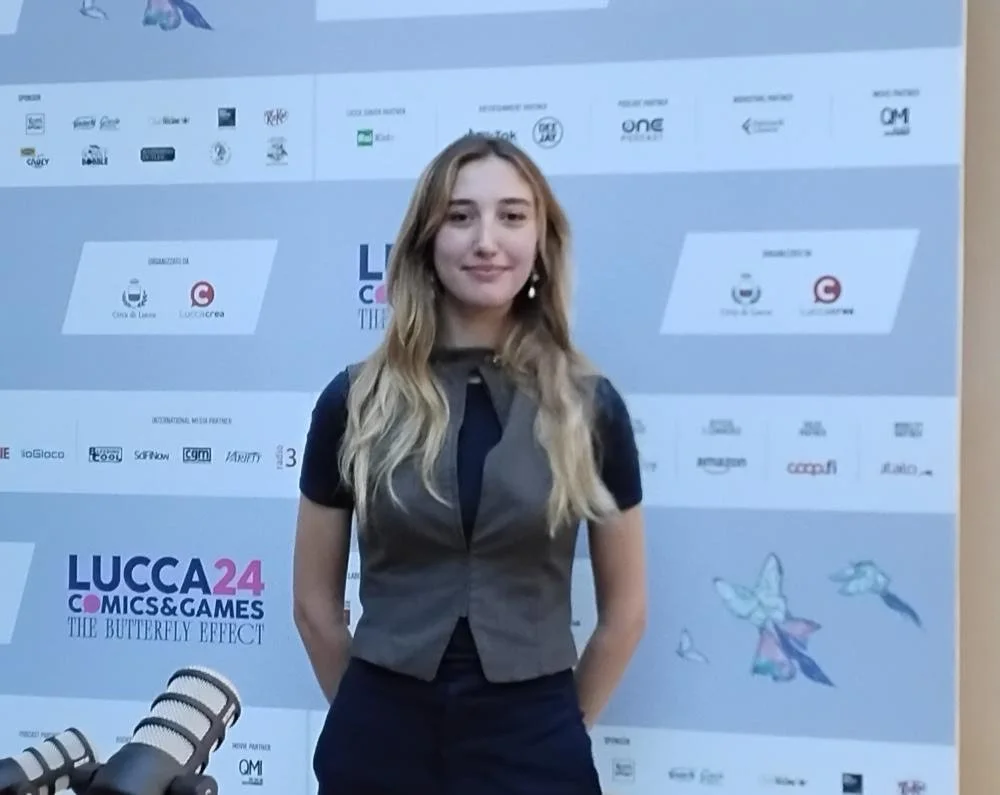 Una giovane donna con capelli lunghi biondi ricci, vestita con camicia nera e gilet grigio, che sorride davanti a uno sfondo con loghi e testo, al backstage di un evento.