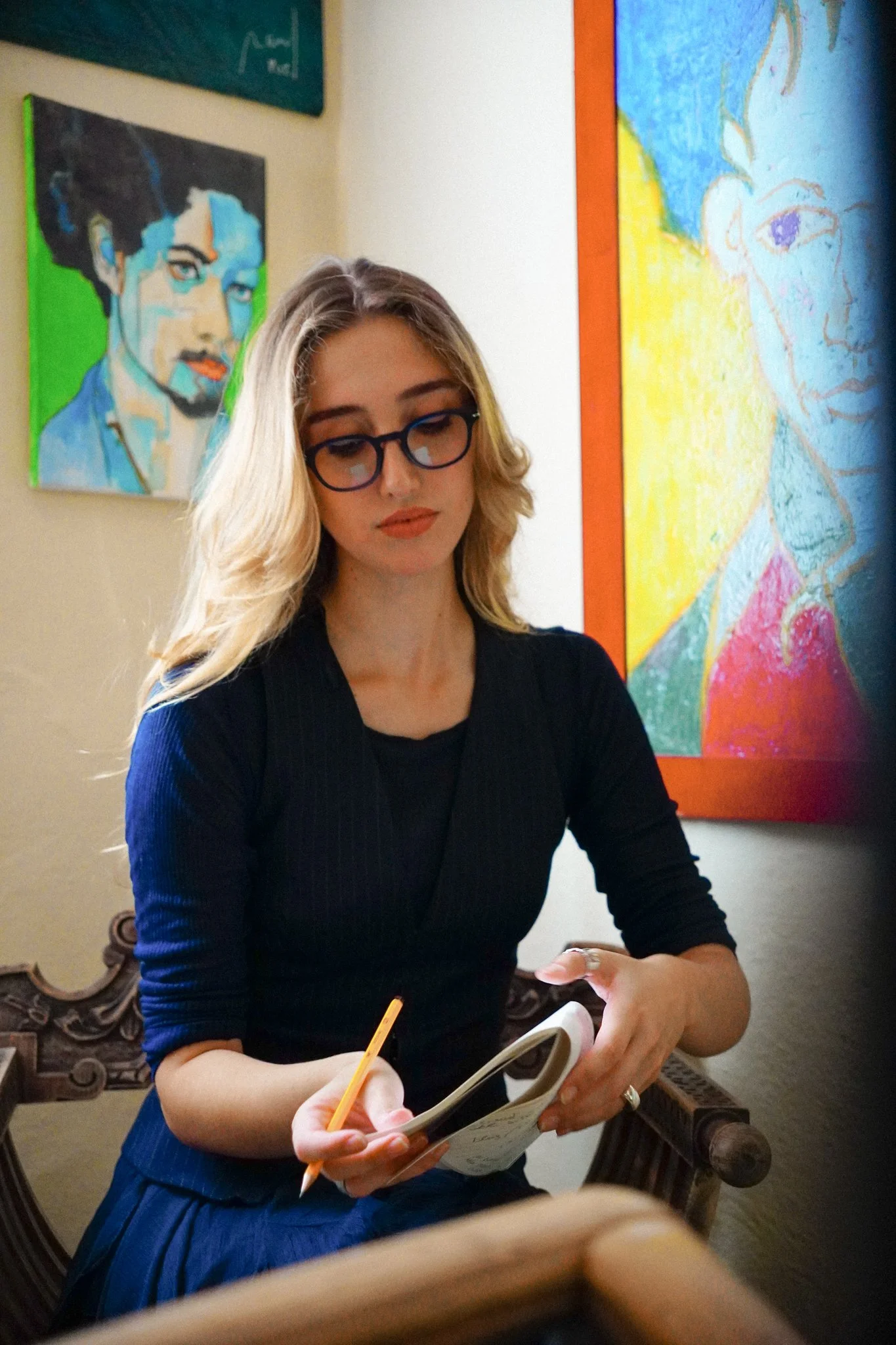 Martina Meroni con occhiali rotondi, abito nero che tiene un libro e scrive con una matita, seduta in un locale con dipinti alle pareti.
