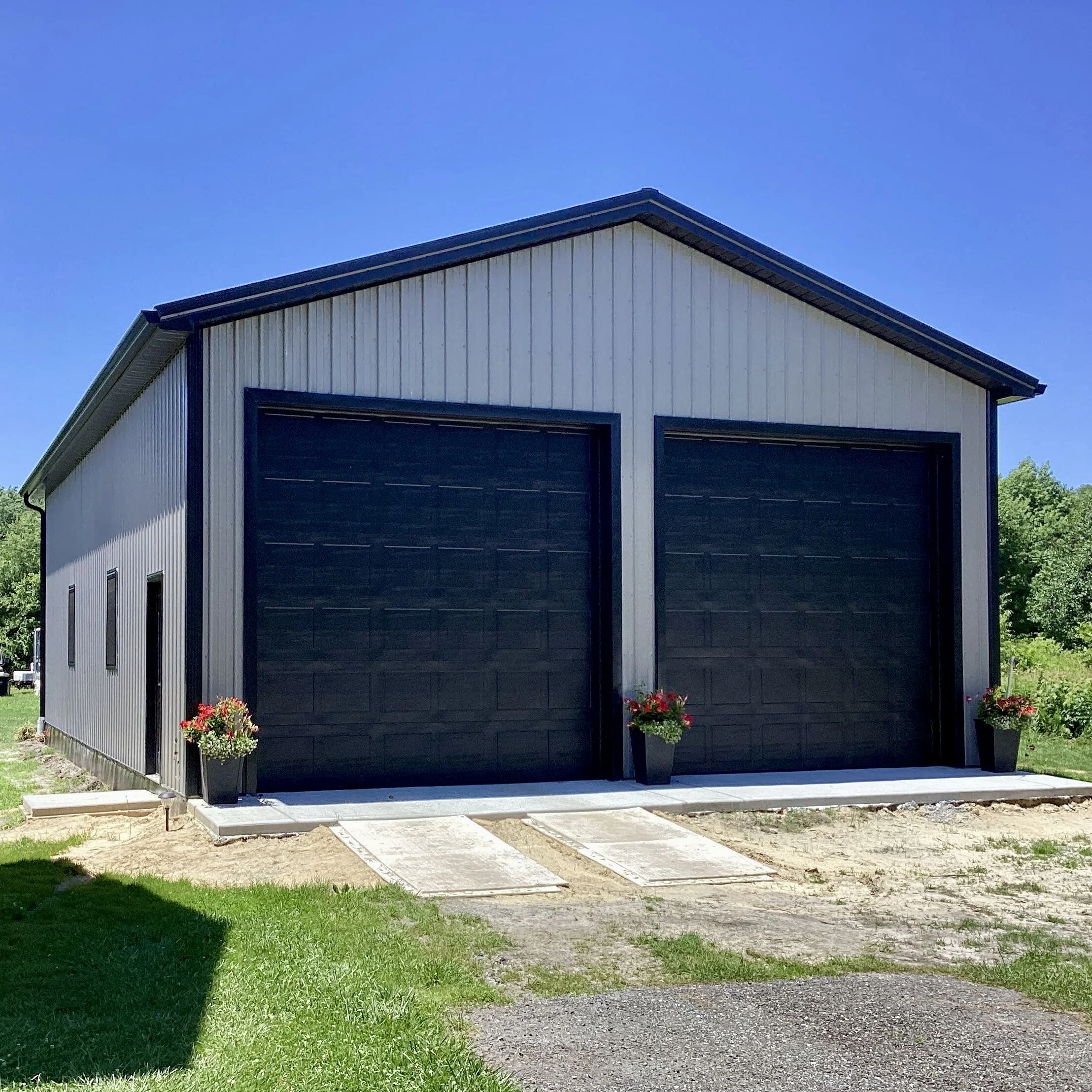 Newly built metal garage with black garage doors, small potted flowers on either side, set against a blue sky, with a gravel driveway and some grass in the yard.