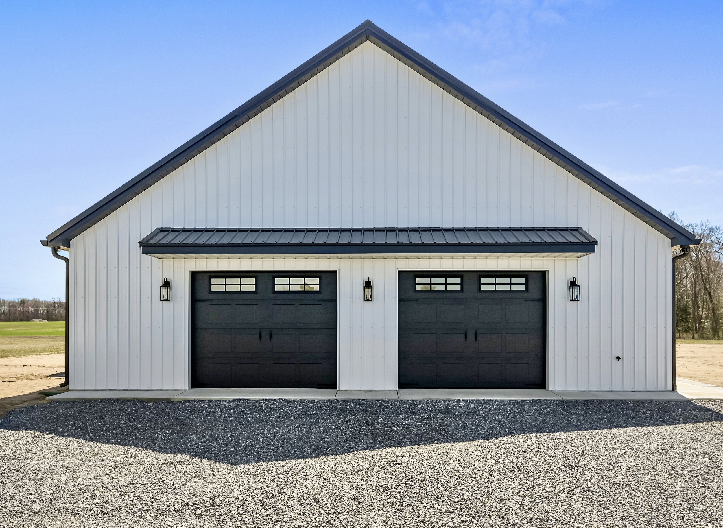 Front view of a white barn with black garage doors and black lantern-style lights, situated on gravel, with a sky background.