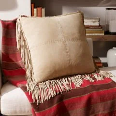 Beige decorative leather pillow with fringe displayed on a red and black plaid textile.