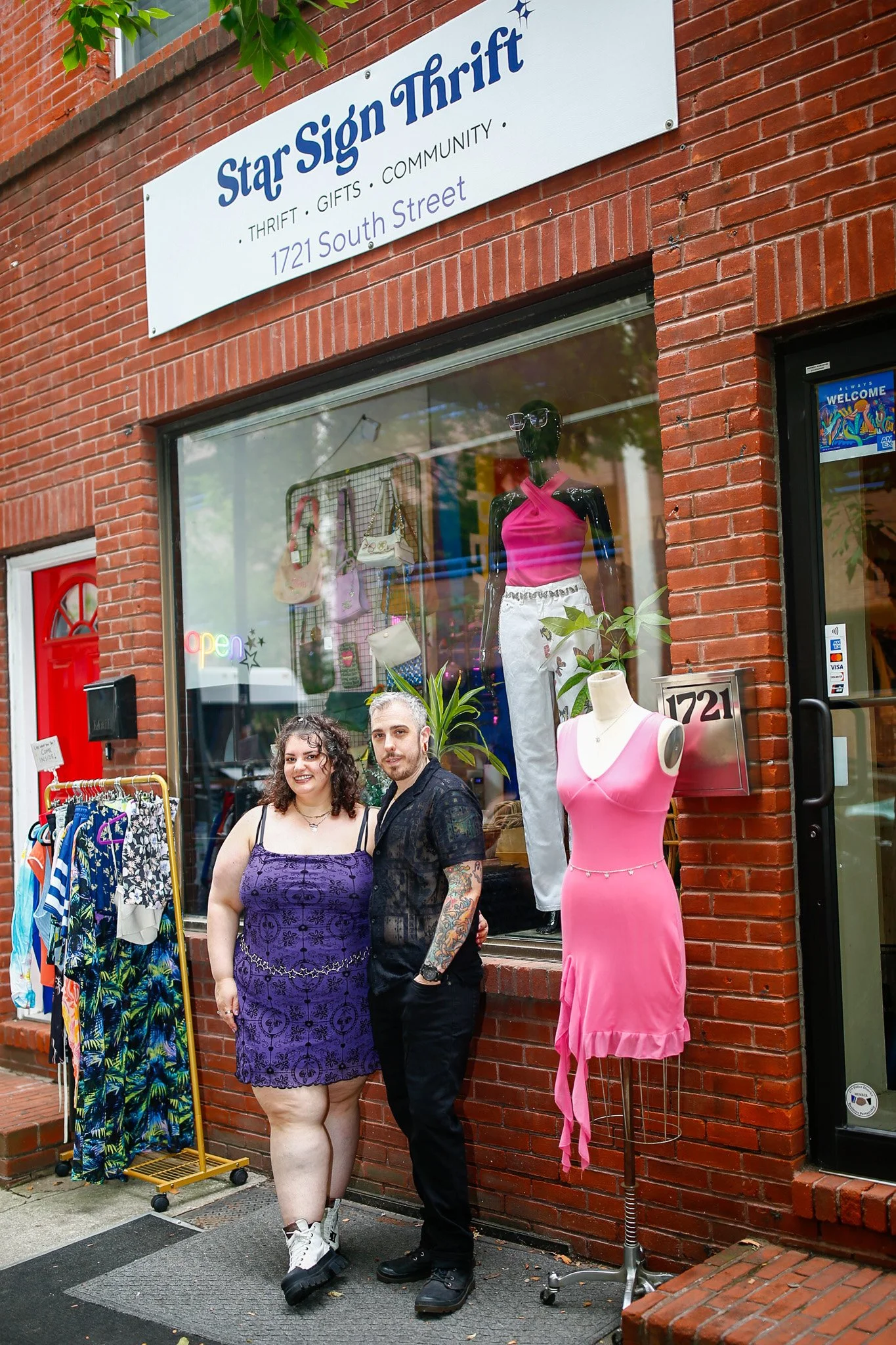 Two people standing outside a clothing store with a brick facade. The woman wears a purple dress with black patterns and sneakers; the man wears black clothing with tattoos visible on his arms. The store window displays mannequins, one in a pink dress and another in white pants and a pink top, along with various bags and clothing items. There is a sign for Star Sign Thrift above the window and a mannequin dressed in a pink halter dress outside.