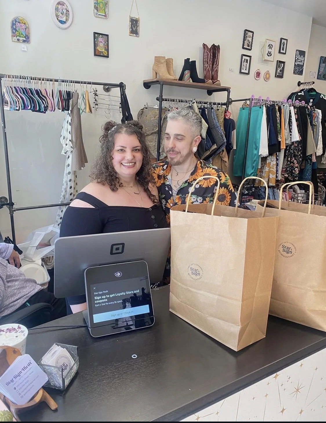 A woman and a man standing behind a checkout counter in a thrift store, smiling at the camera. The store has clothing racks with various garments and wall art, including framed pictures and shoes on shelves.
