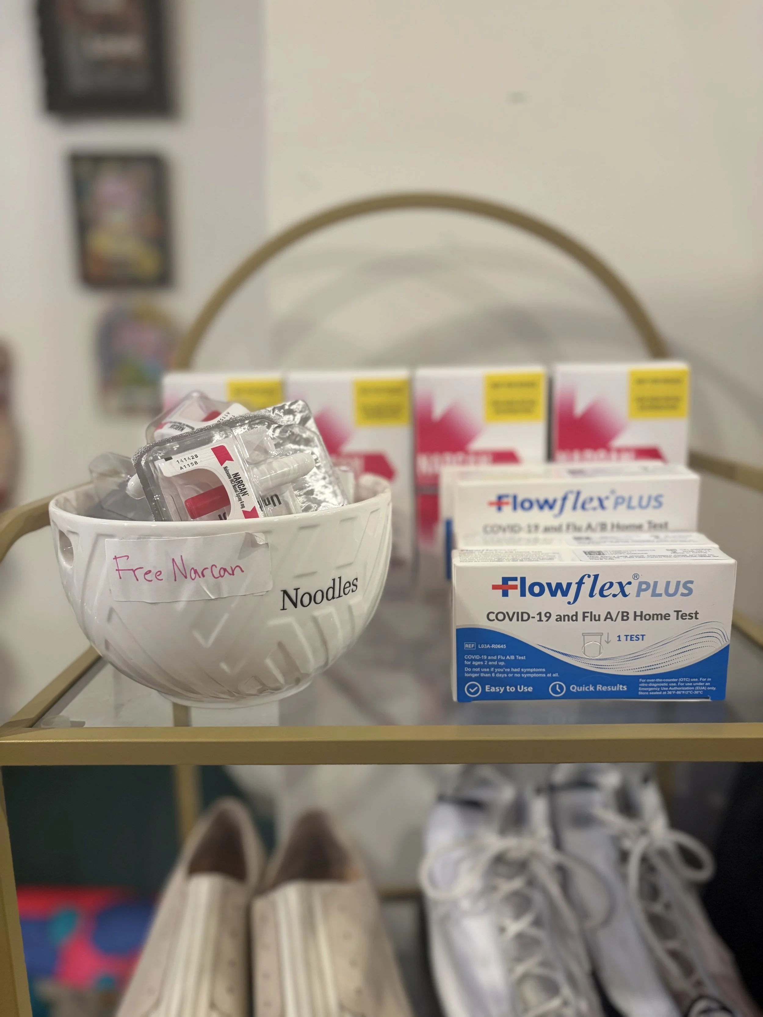 A shelf with medical supplies including COVID-19 and flu home test kits, nasal sprays, and a bowl labeled 'Free Narcan' with nasal spray packages inside.