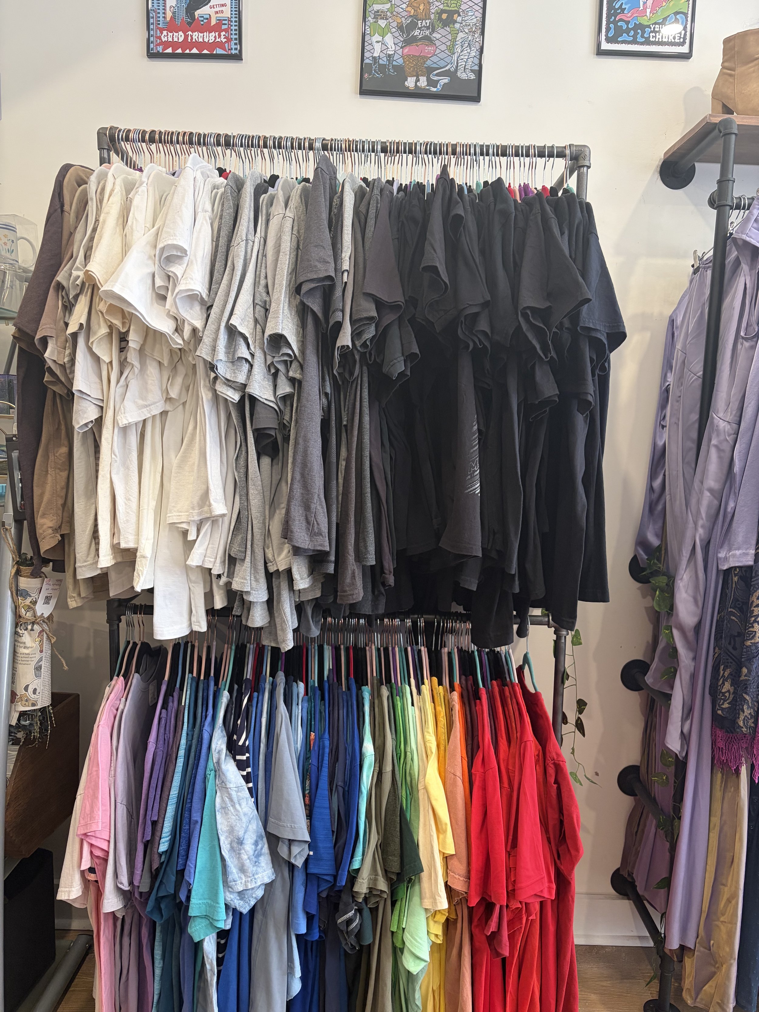 Clothing store display with a clothing rack organized by colors starting from pink, purple, blue, green, yellow, orange, and red. Behind the rack, there are framed comic-style posters on the wall.